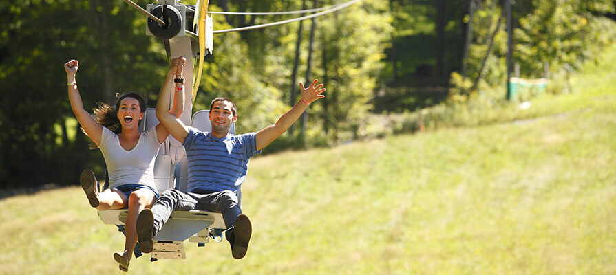 A man and a woman soar with arms up on a ride at Jiminy Peak. They are smiling and happy with green grass and bright sunshine in the background.