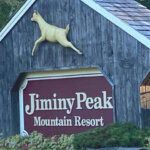 A gray wooden sign for Jiminy Peak Mountain Resort with a golden leaping deer above the maroon sign.