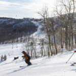 Two skiiers race downhill on a snowy slope at Catamount Mountain Resort with the Berkshire mountains in the background.