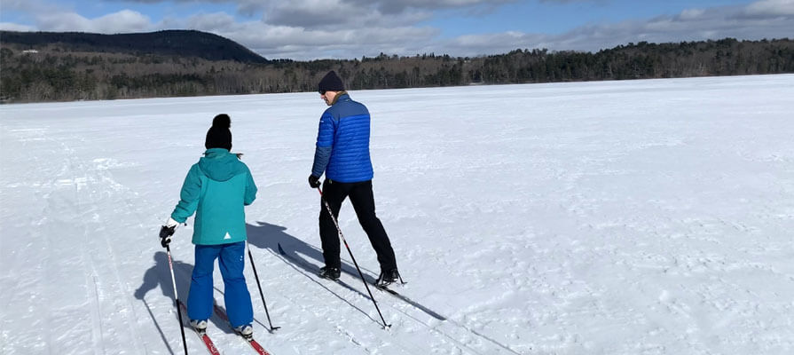 A little girl and father, dressing in snow gear, cross country ski across a frozen lake. The Berkshire mountains are in the distance below a bright blue sky.