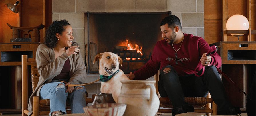 A couple and their yellow lab dog sit in front of a large fireplace at TOURISTS