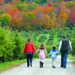 #89 Bartlett’s Orchards, Richmond, MA Photo Credit Kasey Hatch 895×400 Two adults and two children walk hand in hand along gravel path at Bartlett's Orchard with a fall foliage backdrop