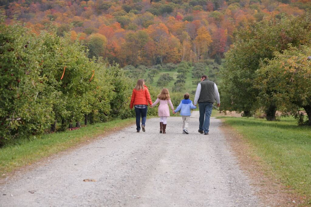 IMG_3551Family at Bartlett Orchard Two adults and two children walk hand in hand along gravel path at Bartlett's Orchard with a fall foliage backdrop.