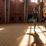 Two people perform yoga poses in the open studio with sun streaming in the windows at Kripalu