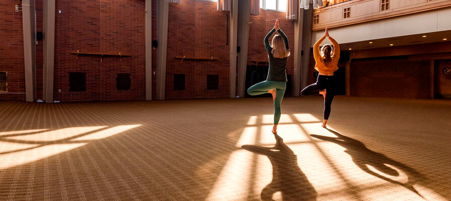 Two women practice yoga in a large room with sunlight shining through the windows at Kripalu Center for Yoga & Health. Photo by Tracy Williams.