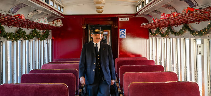 A conductor walks through the festively lit train car with holiday decor.