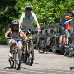 Berkshire Bike and Board_biking outdoors trail (9) 895×400 A family of two adults and a child excitedly ride their bikes on a bright sunny summer day along a paved bike trail.
