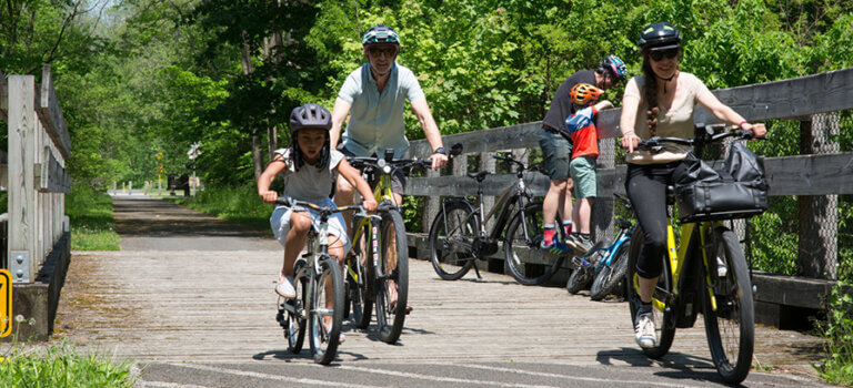 Berkshire Bike and Board_biking outdoors trail (9) 895×400 A family of two adults and a child excitedly ride their bikes on a bright sunny summer day along a paved bike trail.