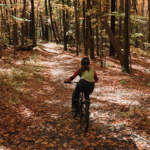 Mountain biking in the fall – credit Great Sky Media (1) 895×400 A woman rides a mountain bike through the shaded forest surrounded by trees and fallen leaves.