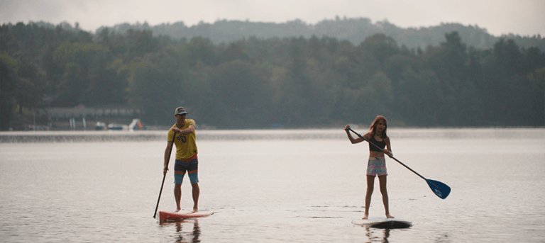 1B Summer 2023_4 Stockbridge Bowl_ paddle boarding_outdoor_water credit Great Sky Media 895×400 A father and daughter paddle board across the Stockbridge Bowl lake on a hazy summer day.