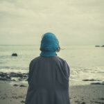 A person in a grey jacket and blue headscarf sits with her back to the viewer along a calm but dusky stretch of beach