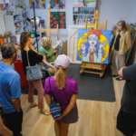 A group of people observing colorful artwork and listening to the artist speak about her work.