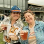 Two smiling women enjoying steins of beer and a large salted pretzel