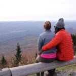 A couple sits on a guard rail overlooking the mountains and fall foliage