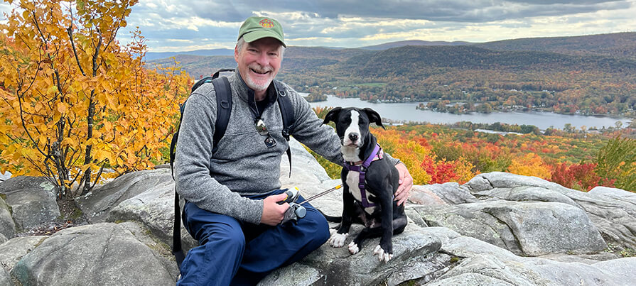 Man and dog smile for photo at the top of a hike surrounded by fall foliage