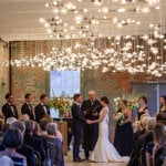Wedding ceremony with bride, groom, and bridal party standing in front of a large gathering at MASS MoCA surrounded by lights across the ceiling