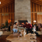 A couple with their dogs cheers their beverages in the lodge at Tourists in front of a cozy fireplace