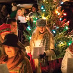 A large crowd of people sing Christmas carols by a decorated tree on a cold night in Stockbridge, MA.