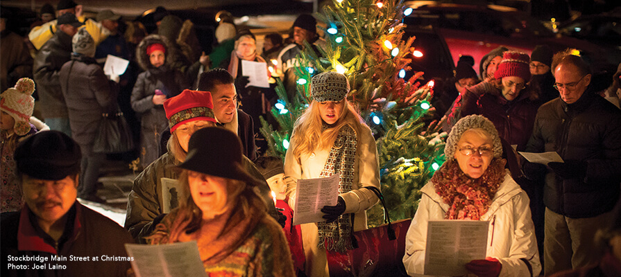 A large crowd of people sing Christmas carols by a decorated tree on a cold night in Stockbridge, MA.