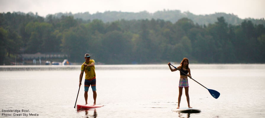 A man and young girl stand on paddleboards in Stockbridge Bowl, a lake in the Berkshires. Green trees line the shore behind them.