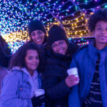 A family of color poses with hot chocolate underneath a tunnel of Christmas lights at Winterlights at Naumkeag.