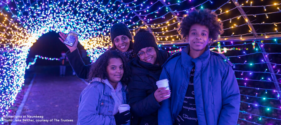 A family of color poses with hot chocolate underneath a tunnel of Christmas lights at Winterlights at Naumkeag.