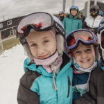 Three kids in ski gear smile at the camera which is using a fish eye lens to distort the image. In the background are smiling adults and Bousquet Mountain ski resort.