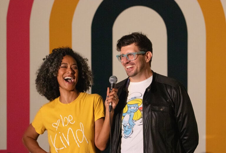 A woman of color in a bright yellow t-shirt that says Be Kind on the front smiles and holds a mic toward the man next to her. The man is white and wears thick rimmed glasses, a white t-shirt, and black leather jacket. He is also smiling.