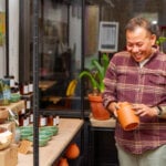 A man of color wearing a plaid shirt smiles and holds a terracotta pot. He is standing in a florist shop. There is a table of pottery and and plants. Behind are large windows. Outside it's night.
