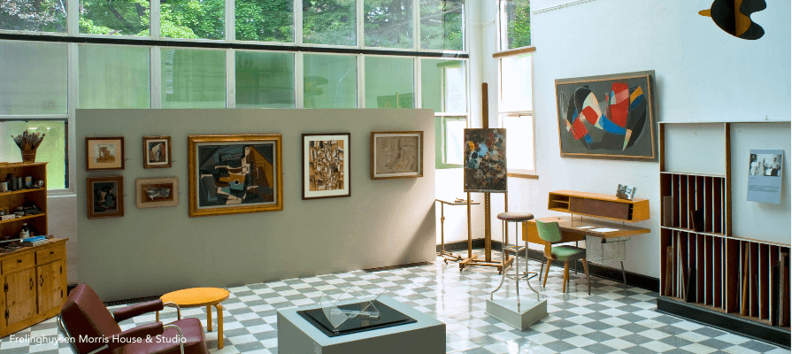 Frelinghuysen Morris House and Studio. This shot shows the interior, a room full of paintings, an easel, chairs, desk, and checkered floor.