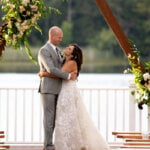 A white man and woman smile at each other beneath wooden beams decorated with flowers. It is a wedding alter. She wears a long lacy white dress. He wears and light gray suit. She has dark brown hair falling down her back. He is bald. Behind them is out of focus greenery.