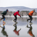 three people in brightly colored jackets skate across the ice with snow capped mountains in the background
