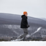 Man in bright orange hat and winter gear snowshoes across a snowy landscape with mountains in the background
