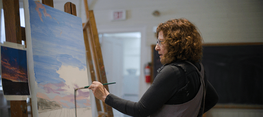 A woman sits in front of a canvas painting a vista scene at the Berkshire Art Center