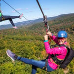 Two women soar along a zipline in the air, wearing bright pink and green jackets, with smiles on their faces in the sunshine