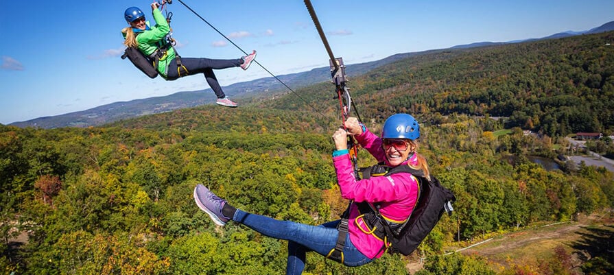 Two women soar along a zipline in the air, wearing bright pink and green jackets, with smiles on their faces in the sunshine