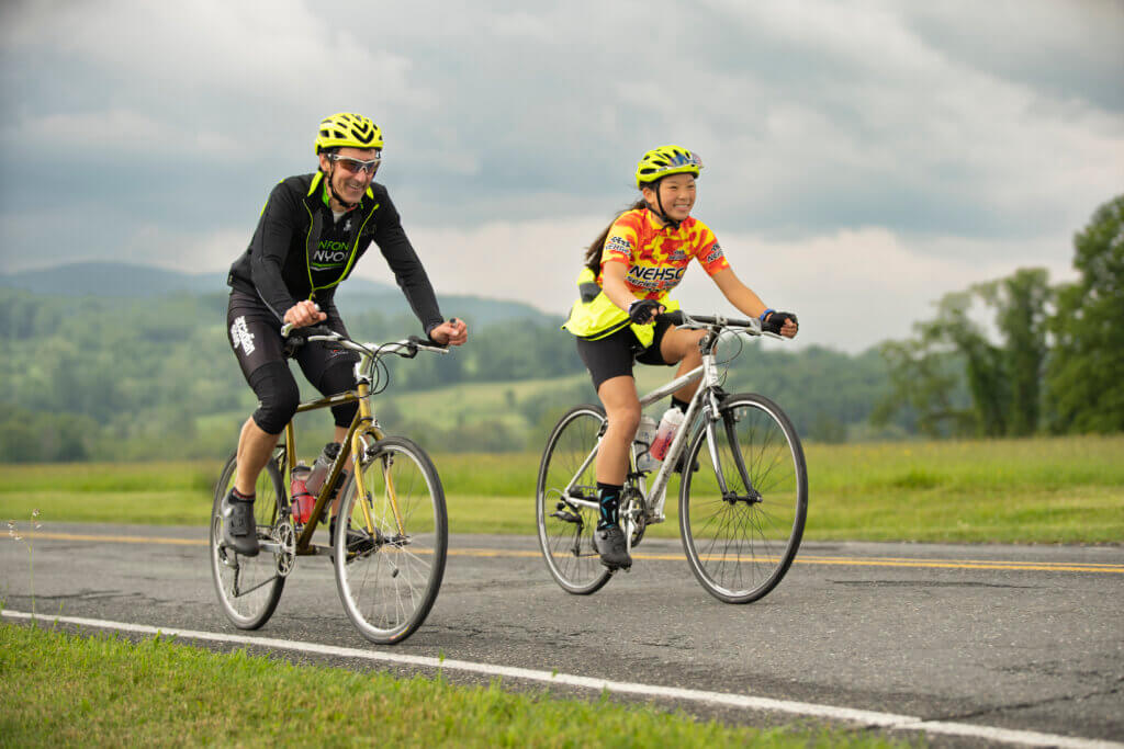 A man and a woman ride bikes roadside on a cloudy summer day.
