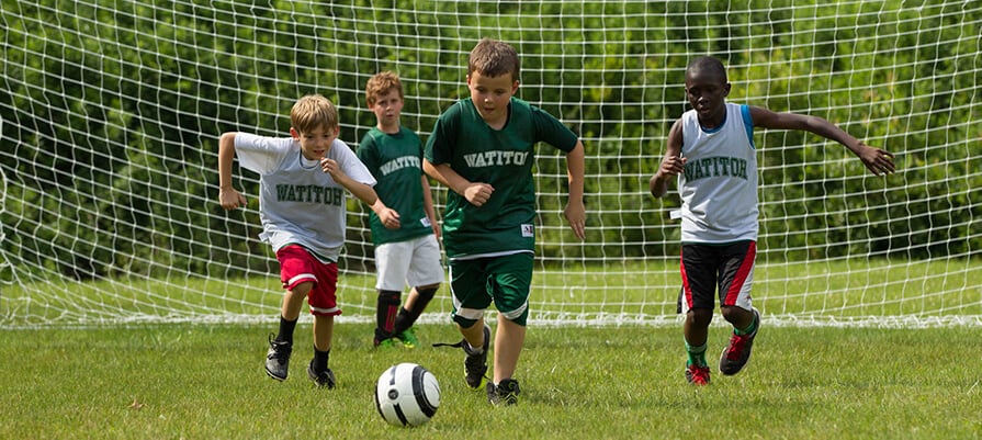 A group of boys chase after a soccer ball on a soccer field at camp Watitoh on a sunny day