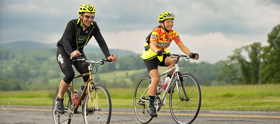 A man and a woman ahppily ride bicycles on an open road along a cloudy skyline.