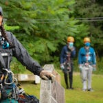 Rachel Hailey, founder of DEI Outdoors, stands on a zipline platform with full harness gear on, in front of a group outdoors