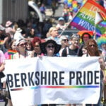 A parade of people walk down the street during the Berkshire Pride Festival, holding a berkshire pride banner with rainbow flags in the background.