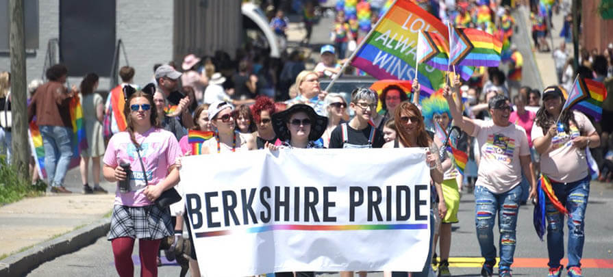A parade of people walk down the street during the Berkshire Pride Festival, holding a berkshire pride banner with rainbow flags in the background.