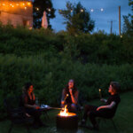 Three people sit in the dark around a fire pit laughing and roasting marshmellows. They are surrounded by greenery, string lights, and a porch above.
