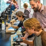 A group of students in a lab look into microscopes with the help of two adults.