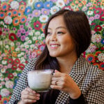 A woman sits in front of a colorful floral backdrop with a large mug in her hands.