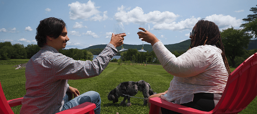 A couple sits looking out over the lawn at Balderdash Cellars. They raise their wine glasses to cheers as a dog sniffs the ground at their feet. 