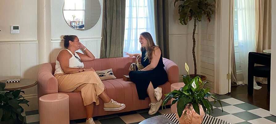 Two women sit in a boutique hotel chatting on a colorful couch. 