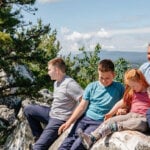 A family of five people sit on a rocky ledge overlooking a beautiful clear scenic view, after hiking.