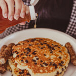 A stack of chocolate chip pancakes sit on a plate with sausage links with fresh maple syrup from Ioka Valley Farm.