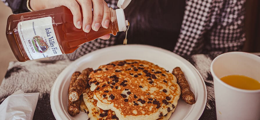 A stack of chocolate chip pancakes sit on a plate with sausage links with fresh maple syrup from Ioka Valley Farm. 